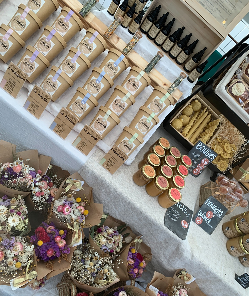 Vu de dessus d'une table au sein d'un marché sur laquelle sont disposés des produits de Lucette et camomille (miel, thé, fleurs, etc)